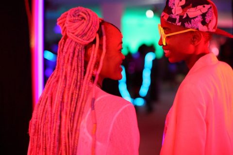 A still from Rafiki. Two people gaze at each other lit by pink light. The person on the left has long braided hair. The person on the right wears sunglasses and a backwards cap.