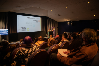 A well lit cinema scene, with a presentation on the screen. people are sat watching one seat apart, wearing masks. a person stands at a podium, we look on as if sat in the back row. over the backs of heads they stand lit, commanding the room.