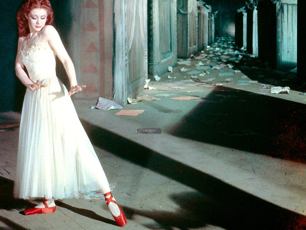 Woman in a shadowy hallway holds a dancelike pose. She wears a white dress and red ballet pumps.