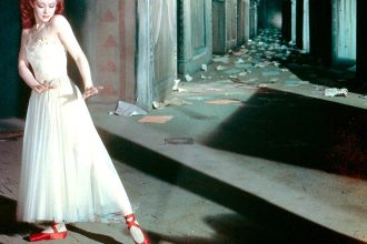Woman in a shadowy hallway holds a dancelike pose. She wears a white dress and red ballet pumps.