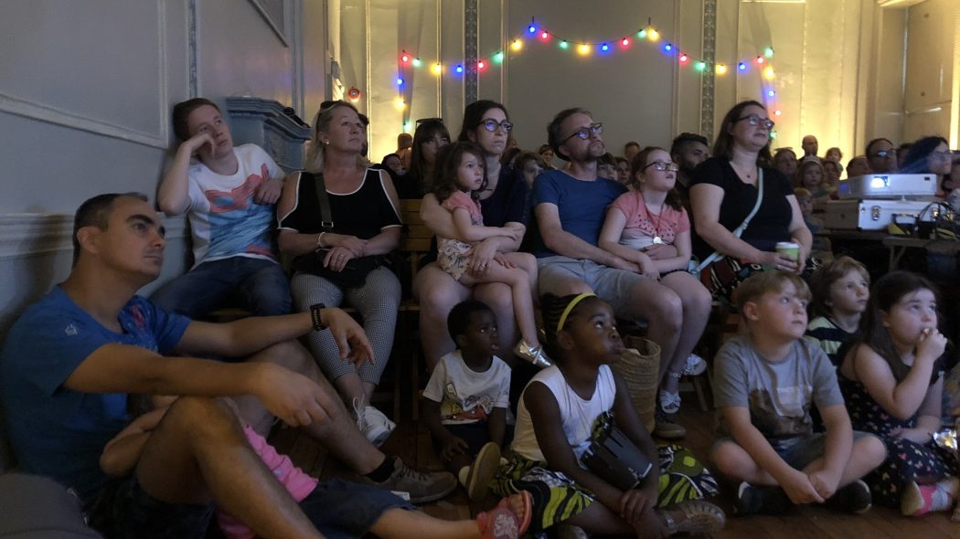 Family audience sit on chairs and the floor looking at a screen behind the camera. Behind them is a string of coloured lights.