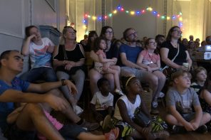 Family audience sit on chairs and the floor looking at a screen behind the camera. Behind them is a string of coloured lights.