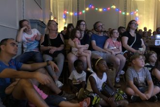 Family audience sit on chairs and the floor looking at a screen behind the camera. Behind them is a string of coloured lights.