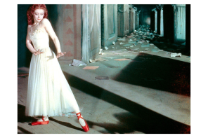 Woman in a shadowy hallway holds a dancelike pose. She wears a white dress and red ballet pumps.