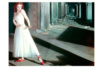 Woman in a shadowy hallway holds a dancelike pose. She wears a white dress and red ballet pumps.