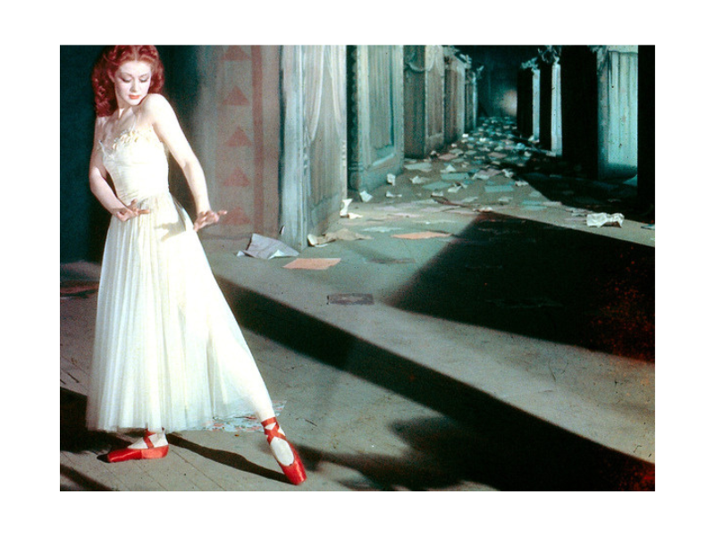 Woman in a shadowy hallway holds a dancelike pose. She wears a white dress and red ballet pumps.